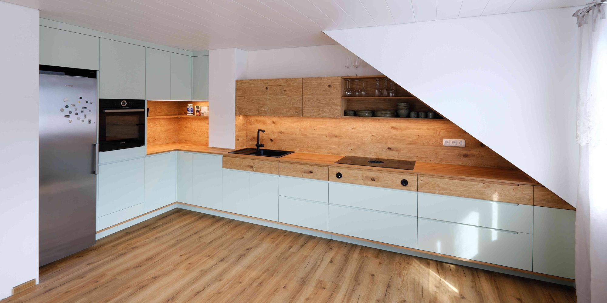 Moderne L-Küche mit mintfarbenen Fronten unter Dachschräge, Eichenholz-Rückwand und LED-Licht