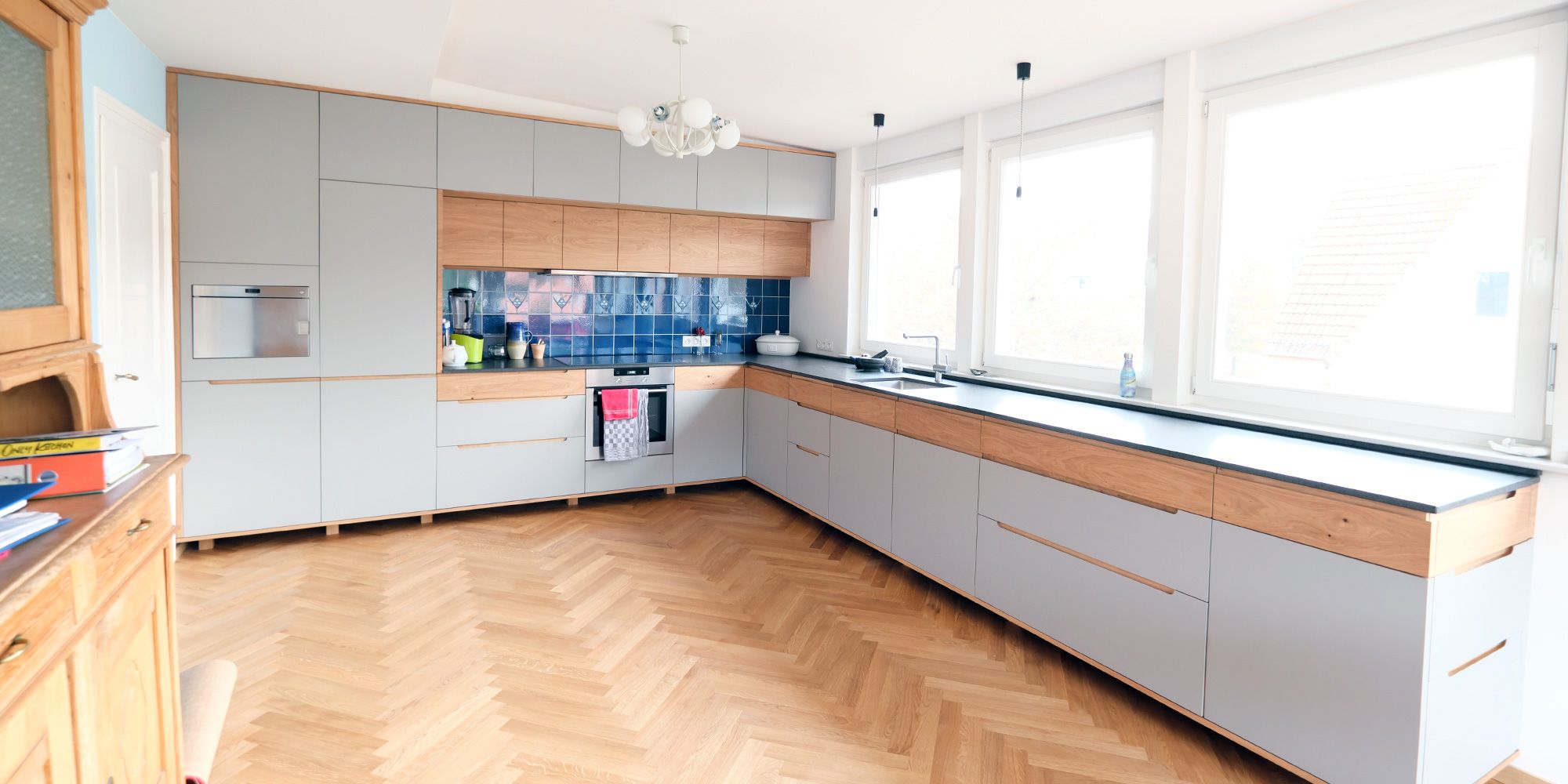 Graue L-Küche mit Eiche-Griffleisten, Fischgrätparkett und blauem Fliesenspiegel unter großen Fenstern