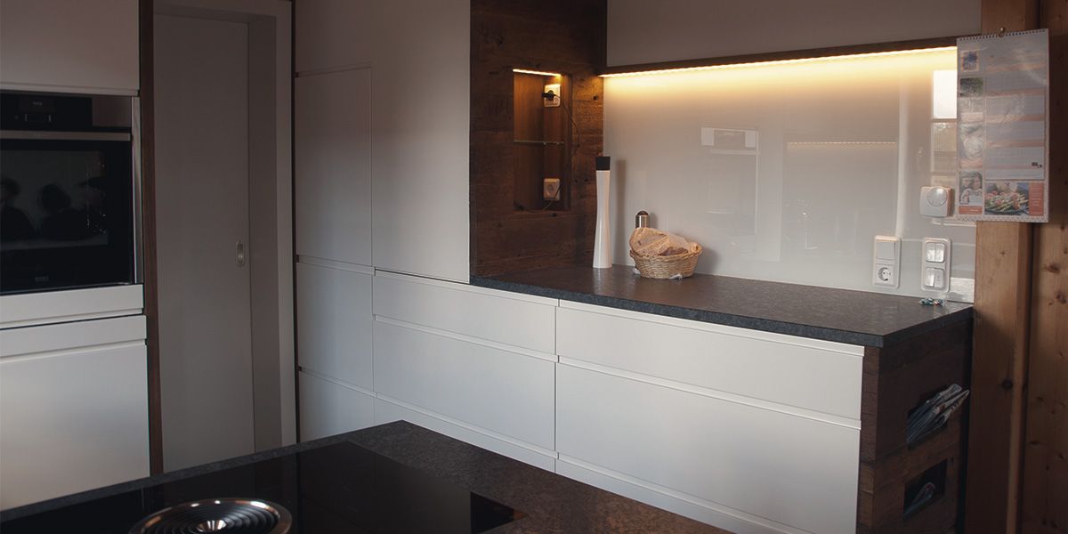 Küchenzeile mit weißen Fronten, LED-Unterbauleuchte und Altholz-Wandverkleidung im warmen Licht