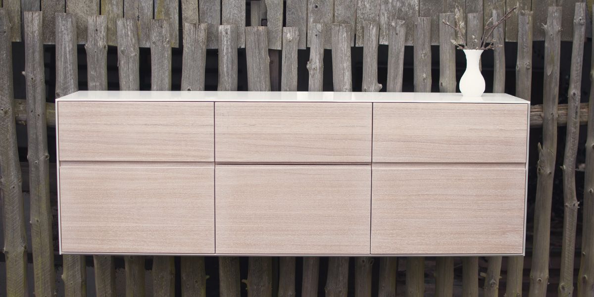 Grifflos wandhängendes Sideboard in Hellholz-Optik vor rustikalem Holzzaun, mit weißer Vase dekoriert