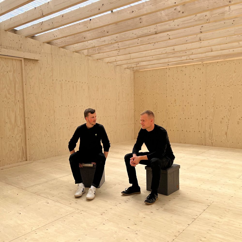 Julian Lauth und Raphael Schneider sitzen im Gespräch in einem holzverkleideten Rohbauraum