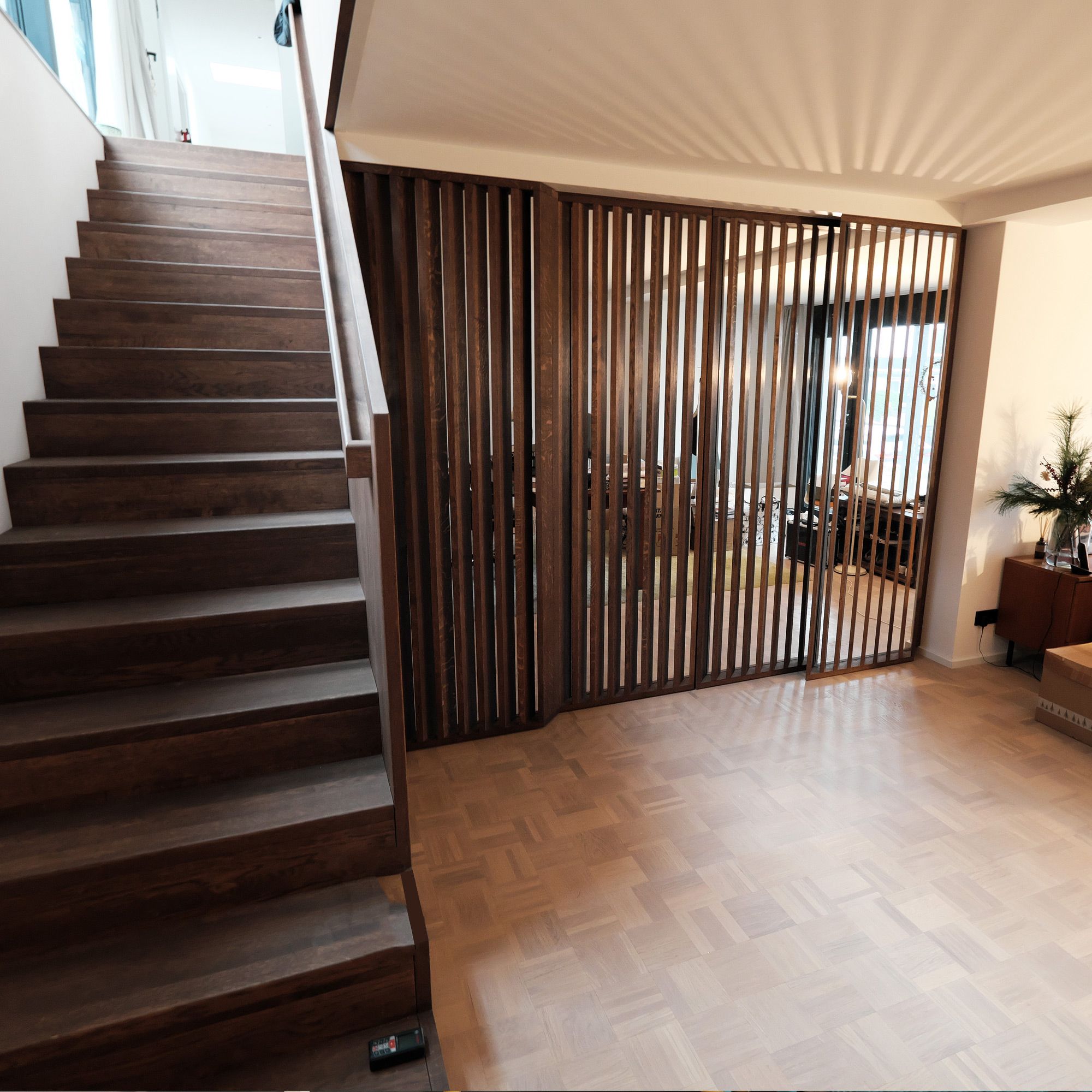 Massivholztreppe mit dunkel geölter Lamellenwand als Raumteiler und Glasschiebetür zum Wohnbereich