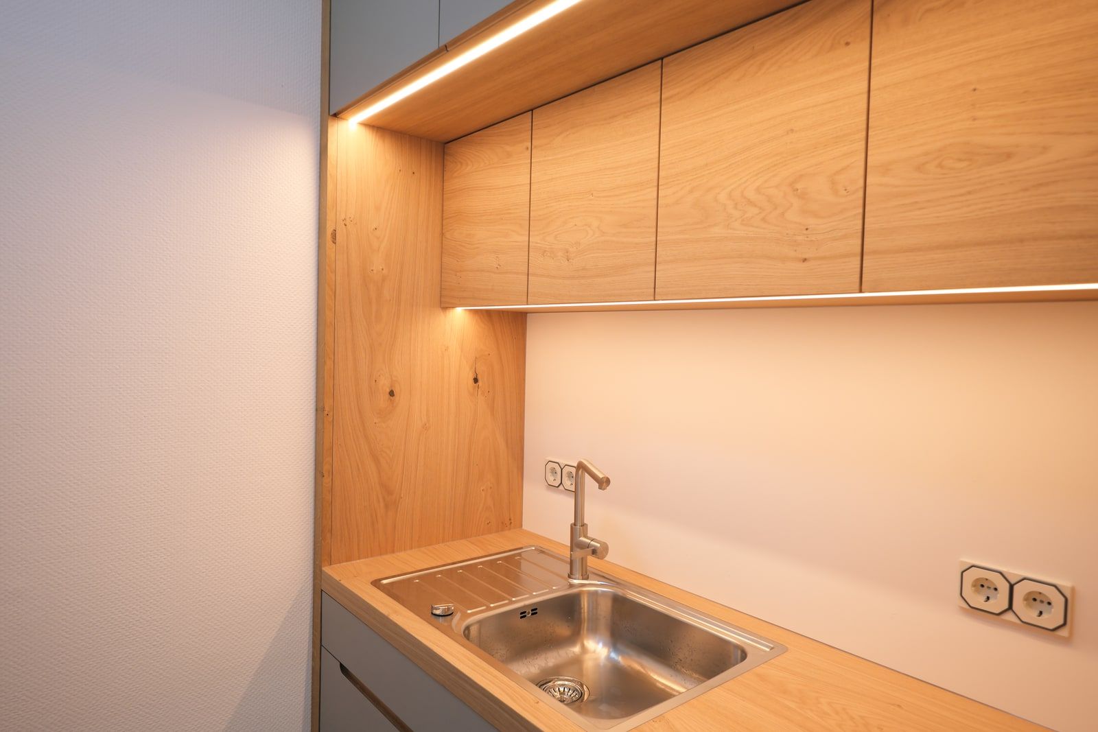 Holzschränke mit LED-Lichtleiste über Edelstahlspüle mit Armatur, warme Lichtstimmung