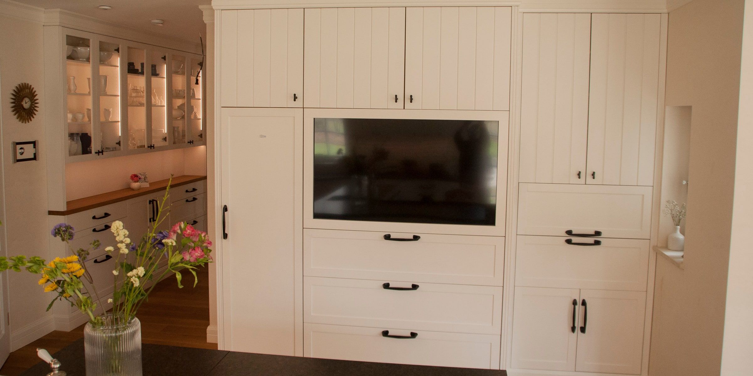 Weiße Landhausküche mit eingebautem TV-Schrank und Kücheninsel mit dunkler Arbeitsplatte