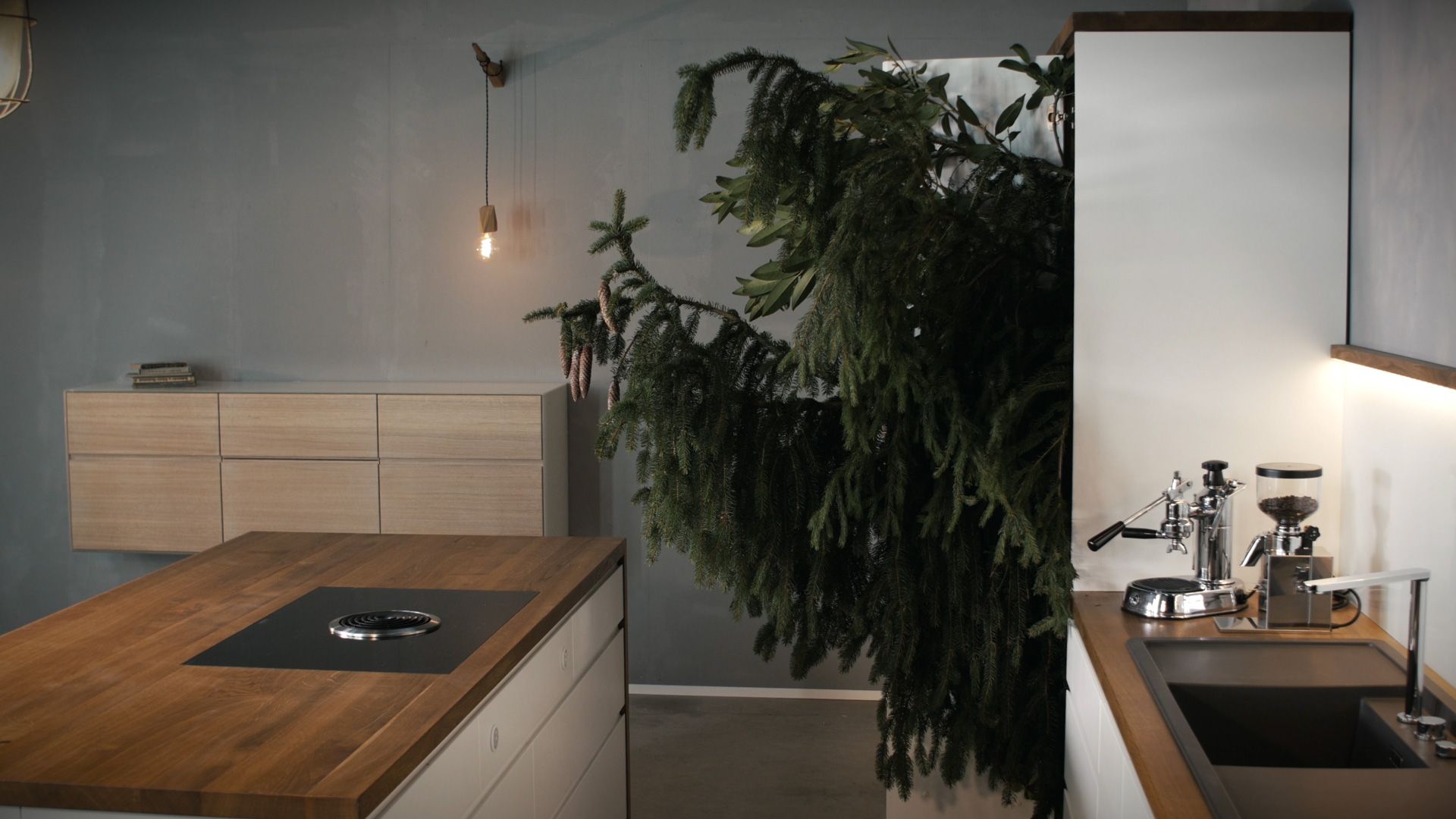 Küche mit Eichenarbeitsplatte, Werkstattlampe und Tannenzweigen vor Betonwand, Szene aus dem Holzfreude-Film